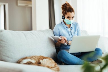 Young woman with protective mask using laptop and writing notes while following classes online from home. 
