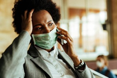 Distraught black businesswoman talking on mobile phone while wearing protective face mask at railroad station hallway. 