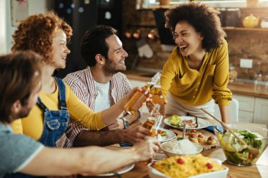 Young happy people having fun and toasting with beer while eating lunch in dining room at home. 