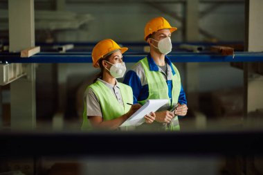 Female worker writing notes while checking stock with her coworker at distribution warehouse during COVID-19 pandemic. 
