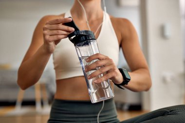 Unrecognizable thirsty athletic woman using water bottle while exercising in the living room. 