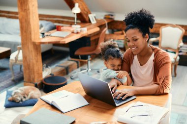 African American stay at home mother working on laptop while daughter is holding her hand and wants to play with her. 