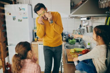 Happy father and his small daughters having fun with vegetables while preparing healthy food in the kitchen.  