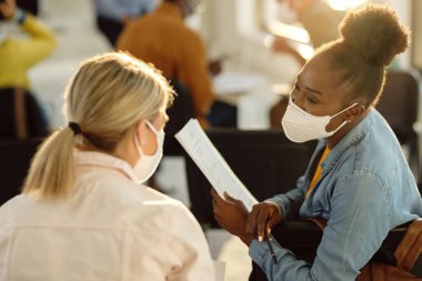 African American college student wearing protective face mask and talking to her friend during a lecture in the classroom. 