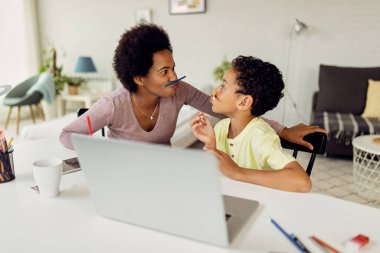 Happy African American mother and son having fun with pencils and making faces while learning at home. 