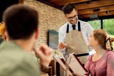 Happy waiter talking to a woman and recommending her what to order from the menu in a restaurant. 