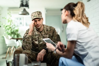 Young military officer holding his head in pain while communicating with healthcare worker who is visiting him at home. 