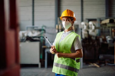 Portrait of female worker holding clipboard and wearing face mask at distribution warehouse due to coronavirus pandemic.