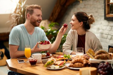 Happy couple having fun while eating breakfast at home. Man is feeding girlfriend with strawberries. 