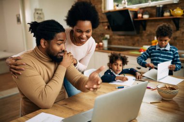 Happy African American woman interrupting her husband who is working on laptop at home. Their children are in the background. 