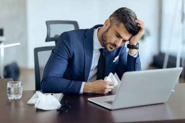 Young businessman feeling unwell and holding his head in pain while working on laptop in the office. 