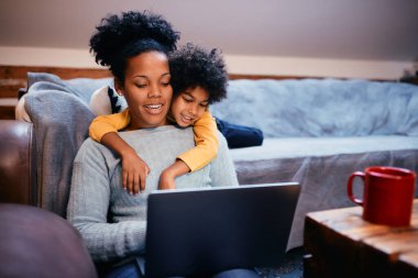 Happy African American mother and daughter surfing the net on laptop while relaxing at home.