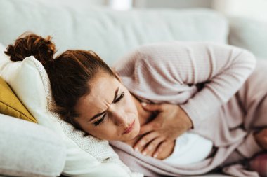 Young woman having chest pain and coughing while lying down on sofa at home. 