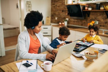 Happy African American single mother working on a computer while the kids are drawing beside her at home. 