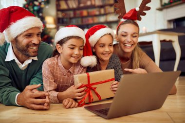 Happy family showing wrapped Christmas present while using laptop and talking to someone via video call at home.