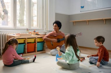 Group of children and their African American teacher playing on instruments during music class at preschool. Focus is on teacher playing acoustic guitar. 