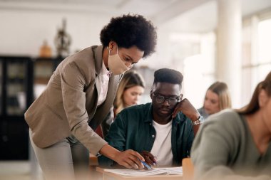 African American professor helping her student with a lecture in the classroom and wearing protective face mask due to coronavirus pandemic.