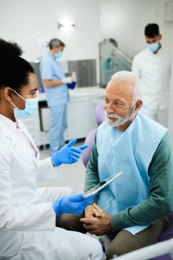 Black female dentist talking to senior patient after dental exam at dentist's office. Focus is on man.