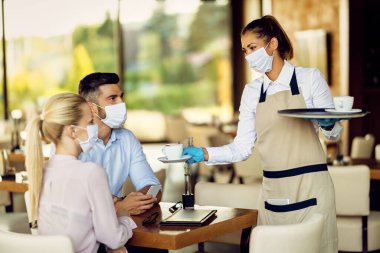 Happy waitress serving coffee to her customers while wearing protective face mask and gloves due to coronavirus epidemic. 