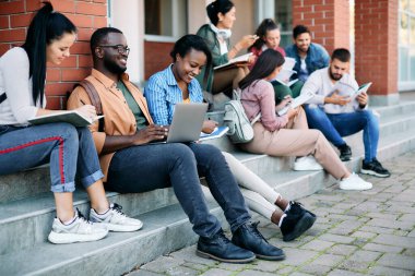 Happy African American student using laptop while studying with female friends in front of university building. 