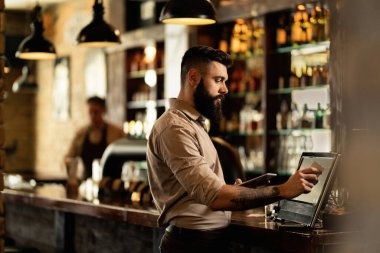 Young bartender using cash register at bar counter in a pub. 