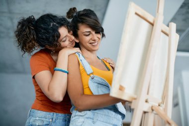 Smiling lesbian hugs her girlfriend who is painting on canvas at home.