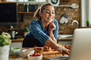 Mutfakta aperatif yaparken bilgisayar kullanan gülümseyen genç bir kadın.. 