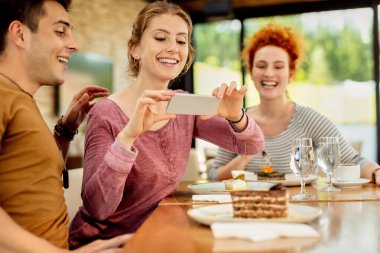 Group of happy friends eating dessert in a cafe. Focus is on woman photographing slice of cake with mobile phone.