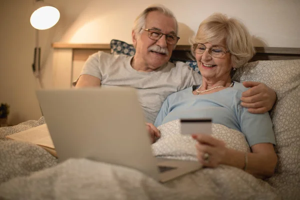 Happy senior couple relaxing in bed while using laptop and credit card for online shopping.