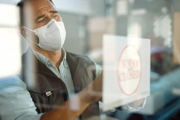 Car repairman with protective face mask hanging closed sign while going out of business due to COVID-19 pandemic. The view is through the glass.