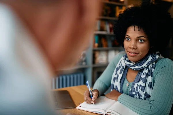 Mid adult African American student writing notes while learning and communicating with her friend in a library.