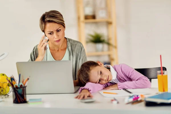 Working mother using laptop and talking on the phone at home. Her daughter is feeling bored while doing homework.