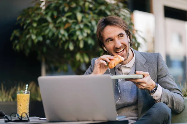 Happy businessman eating croissant while listening music on earphones in outdoor cafe.