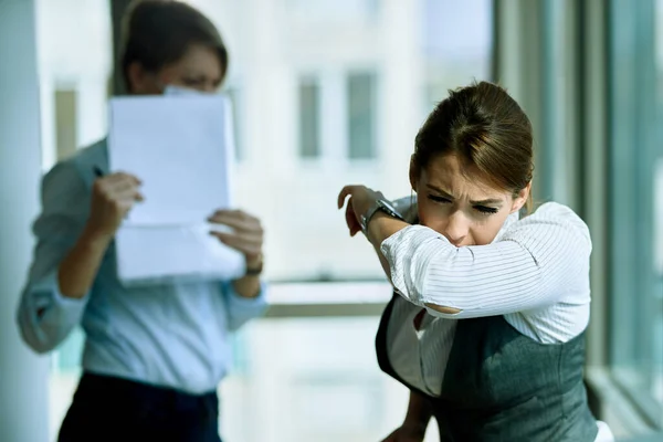 Young businesswoman sneezing into elbow while being in the office. Her colleague is in the background.