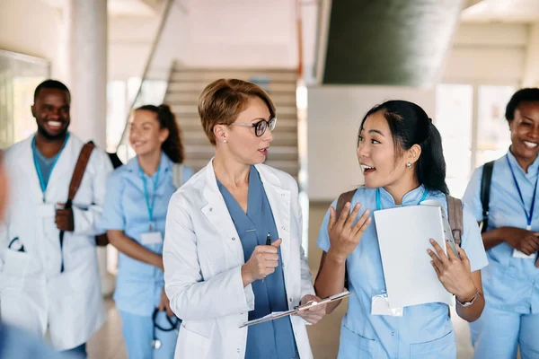 Asian nursing student communicating with female doctor while walking with her friends through hallway at medical university.