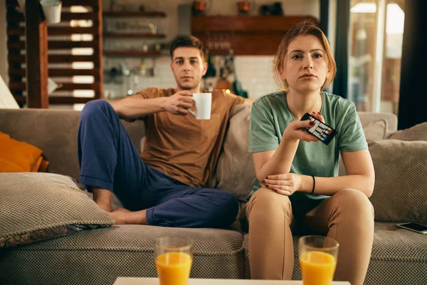 Young woman feeling bored and changing channels while watching TV with her boyfriend at home.