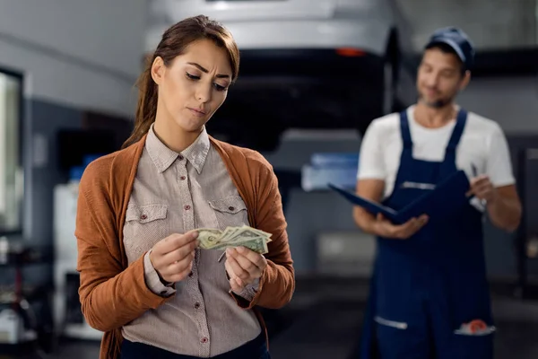 Displeased woman counting money to pay a bill for car repair at workshop.