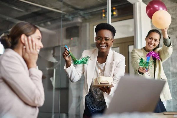 Cheerful female entrepreneurs throwing a surprise party for their colleague Birthday in the office. Focus is on African American businesswoman with a cake.