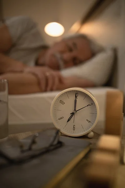 Alarm clock on night table with senior man sleeping in the background.