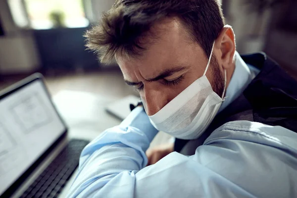 Young entrepreneur coughing into elbow while wearing protective face mask in the office.