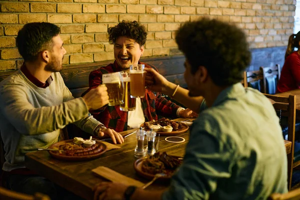 Young man laughing and toasting with beer with his male friends while eating in a pub.