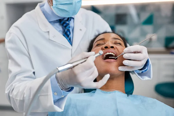 Black woman sitting at dentist\'s chair with eyes closed while dentist is examining her teeth at dental clinic.