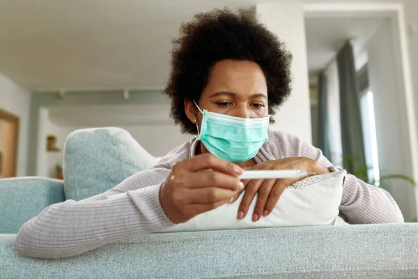 Pensive black woman wearing face mask while measuring her temperature at home.