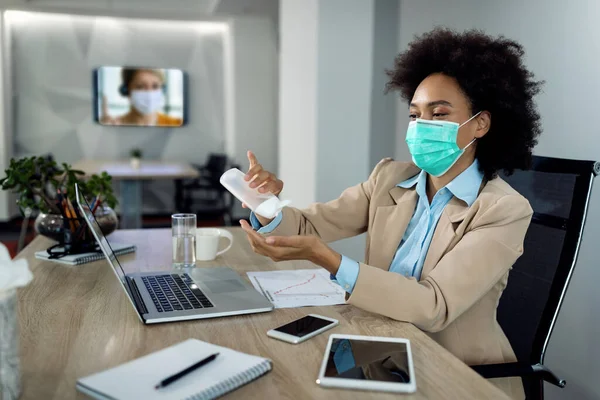 Black businesswoman wearing face mask and disinfecting hands while working in the office during COVID-19 pandemic.
