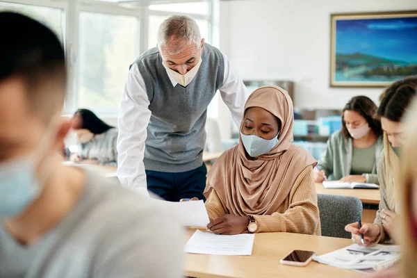 Muslim college student and her teacher analyzing lecture during a class. They are wearing protective face masks due to coronavirus pandemic.