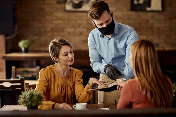 Young waiter serving customers and talking to them while wearing protective face mask in a pub.