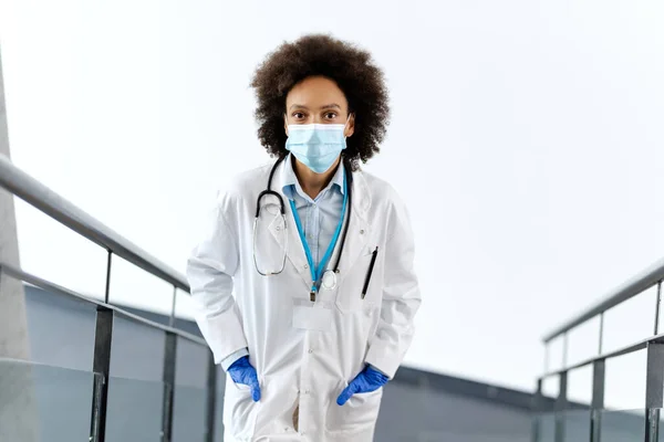 Black female doctor standing at hospital hallway and wearing face mask and gloves due to coronavirus pandemic. Copy space.