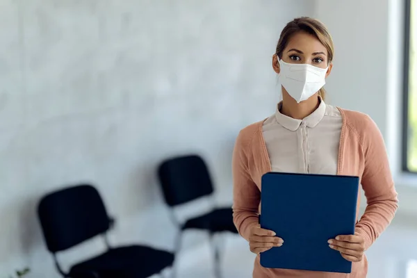 Female candidate wearing protective face mask while waiting for job interview in a hallway and looking at camera.