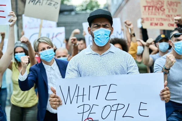 Displeased African American man wearing protective face mask and carrying a placard while protesting with crowd of people on the streets.