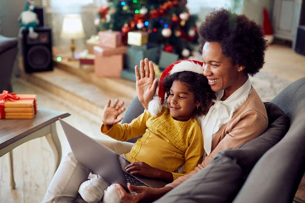 Happy black mother and daughter using laptop and waving during video call on Christmas at home.